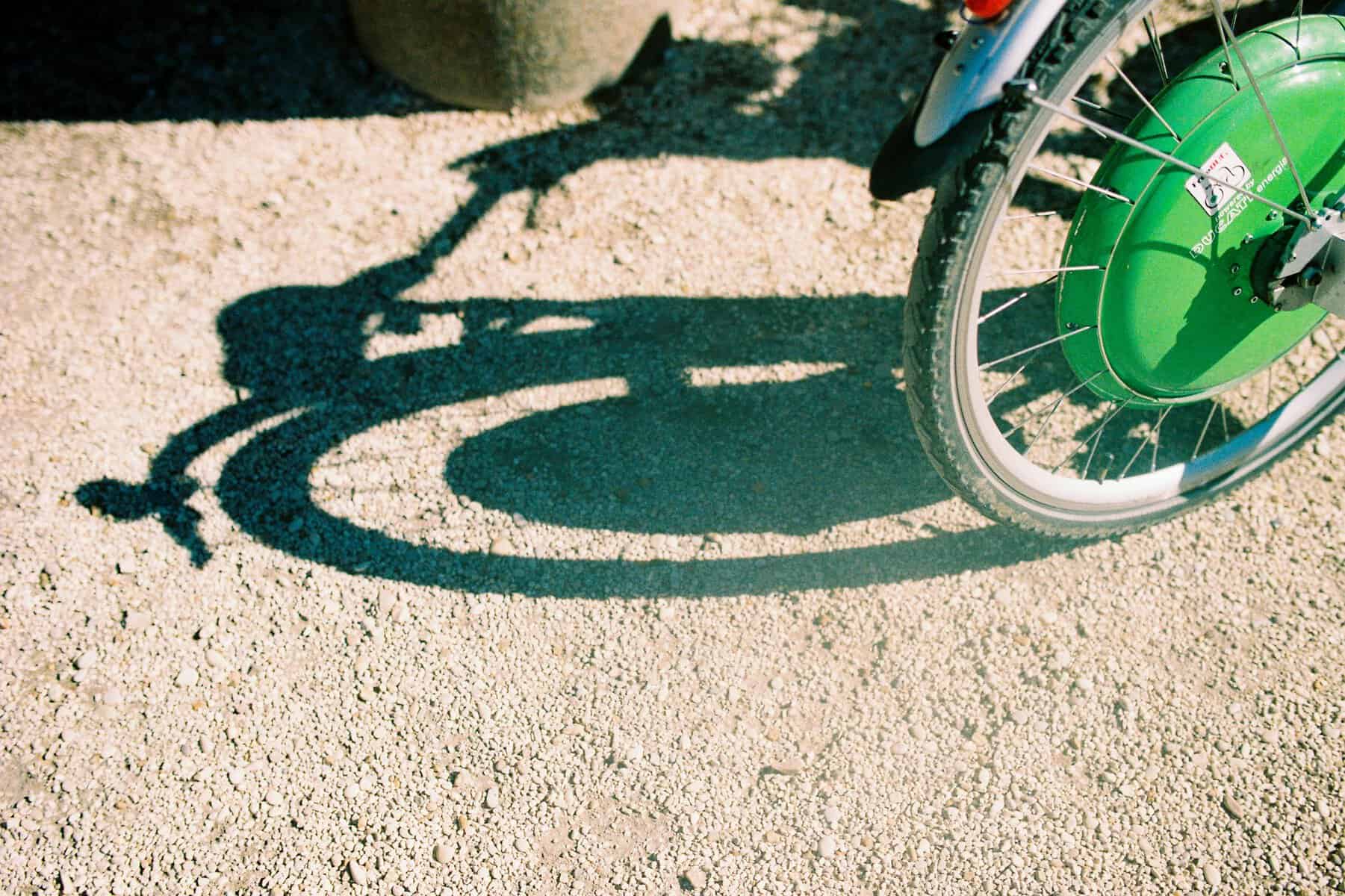 Artistic shadow of a bicycle wheel captured on a gravel path in daylight, highlighting contrast and texture.